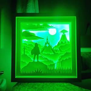May include: A green illuminated papercraft shadow box with a silhouette of a figure holding a sword in front of a castle and a mountain.