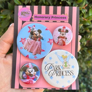 Puede incluir: Un conjunto de cuatro chapas redondas sobre una tarjeta de presentación a rayas negras y rosas con la etiqueta Honorary Princess. Las chapas muestran a Minnie Mouse con un vestido rosa, una Minnie Mouse más pequeña y a Campanilla con el texto Park Princess.