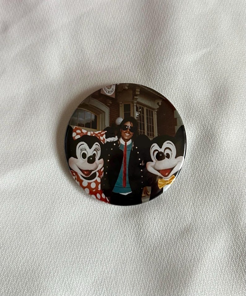 May include: A circular button with a photograph of Michael Jackson posing with Mickey and Minnie Mouse. Jackson is wearing a black jacket and sunglasses. Mickey Mouse wears a yellow bow tie, and Minnie Mouse wears a red polka dot bow.