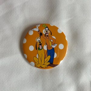 May include: A round button with an orange background and white polka dots. The button features cartoon characters Goofy and Pluto. Goofy is wearing an orange jacket, blue pants, and a green hat. Pluto is yellow with a green collar.