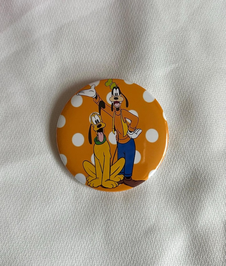 May include: A round button with an orange background and white polka dots. The button features cartoon characters Goofy and Pluto. Goofy is wearing an orange jacket, blue pants, and a green hat. Pluto is yellow with a green collar.