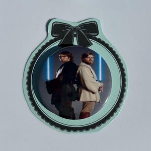 May include: A Star Wars-themed button featuring Anakin Skywalker and Obi-Wan Kenobi with blue lightsabers. The button is set within a light blue scalloped frame with a black bow at the top. The background is white.