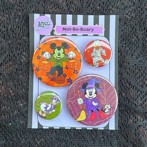 May include: A set of four circular pin-back badges on a display card labelled Not-So-Scary. The badges feature cartoon characters in Halloween costumes, including a mouse with pumpkin buckets, a mummy, a duck, and a mouse dressed as a witch.