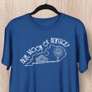 Può includere: Una t-shirt blu reale con una grafica bianca dello stato del Kentucky, una fattoria, una luna crescente e un mulino a vento. Il testo "Blue Moon of Kentucky" è scritto in bianco, con un font usurato attorno al contorno dello stato.