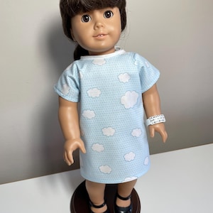May include: A doll wearing a light blue hospital gown with white clouds printed on it. The gown has a white trim around the neckline. The doll is wearing a white hospital bracelet that says "SAMANTHA DOB 12/10/1998".