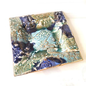 May include: A square ceramic dish featuring a textured surface with a glaze in shades of deep blue, turquoise, and earthy tan. The piece displays various impressed patterns, including cross-hatching, leaf motifs, and organic shapes across its surface.
