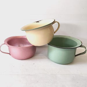 May include: Three vintage enamel chamber pots in pastel shades: pink, pale yellow, and mint green. Each pot has a handle and a contrasting rim. The pots are arranged on a white wooden surface.