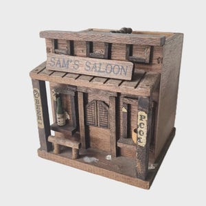 May include: A miniature wooden model of "Sam's Saloon" with a distressed finish. The saloon includes a small bench, a bottle, and signs for "Beer" and "Pool". The building is made of brown wood and has a rustic design.