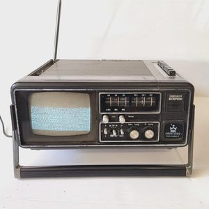May include: A vintage Princess Walkabout portable television and radio combination unit in a dark grey casing. It features a small screen with static, a retractable antenna, a condenser microphone, and various manual tuning knobs and switches.