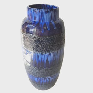 May include: A tall, ceramic vase with a glossy finish. The vase has a dark blue interior and a textured, dark grey band around the middle. The exterior has a dripped blue glaze effect, creating a unique decorative piece.