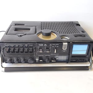 May include: A black portable television and cassette recorder combination unit with a textured leather-like finish. The device features a small screen, a top-loading cassette deck, a speaker grille, multiple control knobs, and switches.