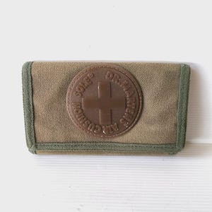 May include: An olive green and tan canvas wallet with a brown leather patch. The patch displays the Dr. Martens logo and a cross. The wallet has a green trim.
