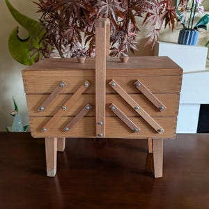 May include: A wooden sewing box with a hinged lid and a carrying handle. The box has three compartments and is decorated with a geometric pattern. The box is sitting on four legs.