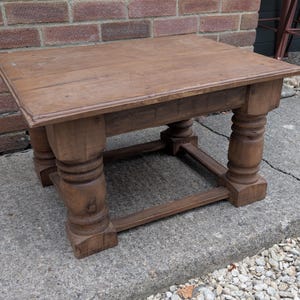 May include: A low, rectangular wooden table with a natural brown finish. The table features a thick, flat top surface supported by four sturdy, turned legs with a rounded, decorative profile. A horizontal wooden stretcher connects the legs.