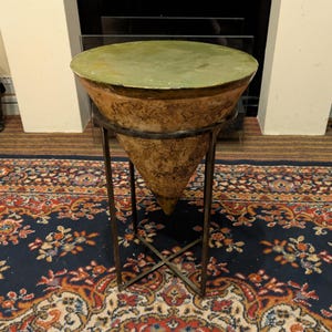 May include: A conical terracotta vessel with a weathered, textured brown exterior and a smooth, flat, olive-green circular top. The vessel is supported by a dark metal stand featuring a circular ring frame and four straight legs with a cross-base.