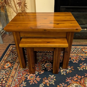 May include: A set of two rectangular nesting tables crafted from polished brown wood with a visible grain pattern. The larger table features a smooth top surface and four sturdy legs, while the smaller table fits underneath it.
