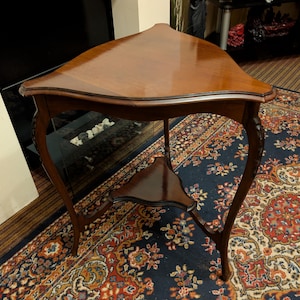 May include: A vintage, three-legged, triangular side table. The table is made of dark wood with a glossy finish. It features a small shelf and ornate, curved legs. The table is approximately 76 cm wide.