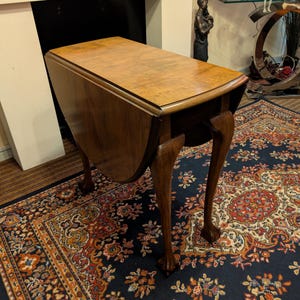 May include: A dark wooden drop-leaf table with a polished finish. The table has a rectangular top with curved drop leaves. The legs are curved with claw feet. The table is set on a patterned rug, and the overall design is classic.