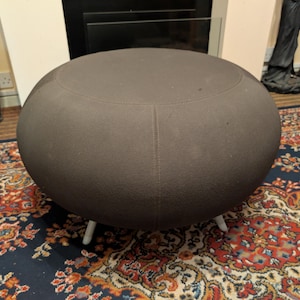 May include: A round, dark brown ottoman with a textured fabric surface and a smooth, curved silhouette. The piece features a flat top and is supported by short, light-coloured legs that elevate the base slightly above the floor surface.
