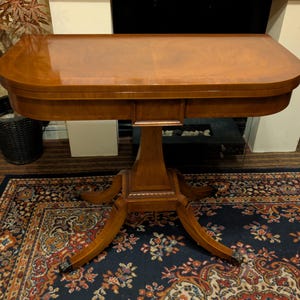 May include: A wooden, oval-shaped table with a glossy finish. The table has a pedestal base with four curved legs. The table is set on a patterned rug. The table is approximately 102 cm long and 51 cm wide.