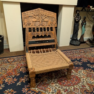 May include: A low-profile wooden chair featuring a hand-carved backrest with two elephants and floral motifs. The seat is constructed from woven natural fibre rope, and the four legs are carved with floral patterns, providing a sturdy base.