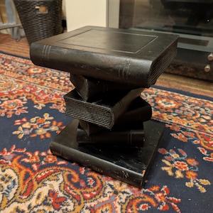 May include: A black side table constructed from a stack of five rectangular books. The base is a large book, with four smaller books stacked in a staggered, rotating pattern to form the central column, topped by a flat, rectangular book surface.