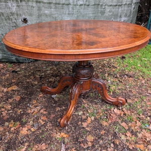 May include: An oval wooden table with a polished burl wood grain top. The table features a central pedestal base with four carved, curved legs that extend outwards, showcasing intricate floral and scrollwork detailing in the dark wood.