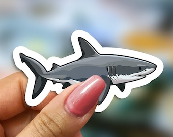May include: A white and gray shark sticker with a white outline. The sticker is being held by a hand.
