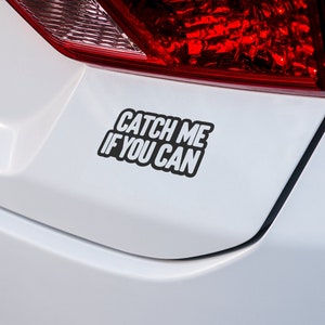 May include: A white car with a black bumper sticker that reads "Catch Me If You Can".