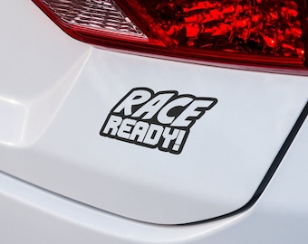 May include: A white car with a black "Race Ready!" sticker on the rear bumper.