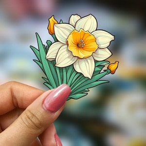 May include: A white and yellow daffodil flower sticker with green leaves. The sticker is held in a hand with pink nail polish.