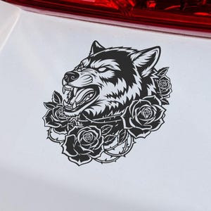 May include: Black and white decal of a snarling wolf's head surrounded by roses. The sticker is affixed to the white surface of a vehicle, near the red taillight.