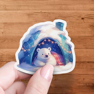 May include: A white polar bear sits inside a blue and white igloo with a red and white blanket. The igloo has a snowy roof and a chimney. There are string lights inside the igloo.