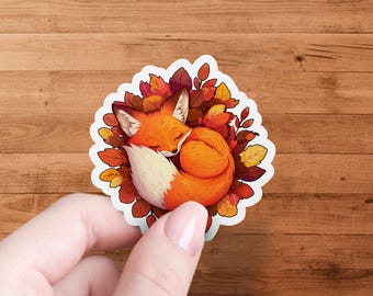 Op de afbeelding: Een sticker met een opgerolde oranje vos die slaapt in een bed van herfstbladeren. De vos is omringd door rode, oranje en gele bladeren.