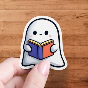 Könnte beinhalten: Ein weißer Geisteraufkleber mit schwarzem Umriss, der ein Buch hält. Das Buch hat einen blauen und einen orangefarbenen Einband mit gelben Seiten. Der Geist hat schwarze Augen und rosa Wangen. Der Aufkleber wird von einer Hand vor einem Holzgrund gehalten.