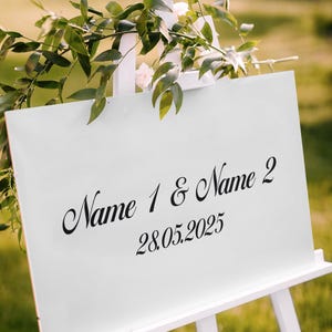 May include: A white sign on a white easel, decorated with greenery and flowers. The sign displays the names "Name 1 & Name 2" and the date "28.05.2025" in elegant black script, perfect for a wedding or event.