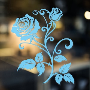 Op de afbeelding: Een lichtblauwe roos met een stengel en bladeren, met een decoratief swirl-ontwerp. De roos bevindt zich op een helder glazen oppervlak.