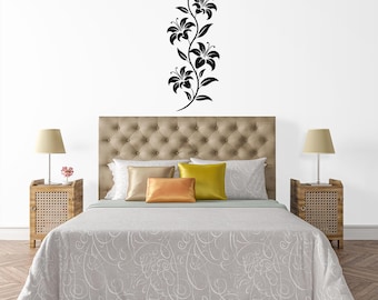 May include: A black floral wall decal featuring lilies, applied above a bed with a tufted headboard. The bed is made with a patterned comforter and decorative pillows. Two bedside tables with lamps flank the bed.