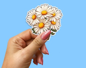May include: A sticker featuring a bouquet of white daisies with yellow centers. The sticker is held in a hand with pink nail polish.