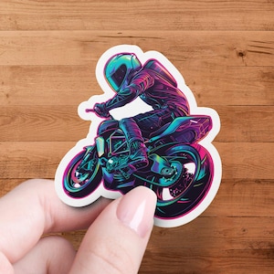 Puede incluir: Una pegatina con una ilustración colorida de una persona conduciendo una motocicleta. La persona lleva un casco y la motocicleta está en el aire. La pegatina está sobre un fondo de madera.
