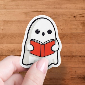 Pode incluir: Um autocolante de fantasma branco com contorno preto, segurando um livro vermelho. O fantasma tem olhos pretos e uma expressão de surpresa. O autocolante é segurado por uma mão sobre um fundo de madeira.