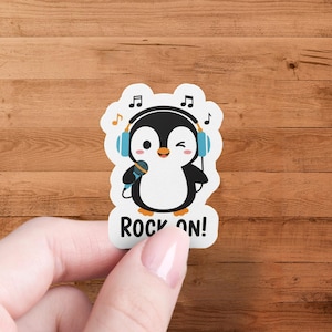 Puede incluir: Una pegatina con un pingüino de dibujos animados que lleva auriculares y sostiene un micrófono. El pingüino es blanco y negro con detalles en azul. La pegatina tiene las palabras "ROCK ON!" en la parte inferior y notas musicales alrededor del pingüino.