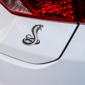 May include: Black and white cobra emblem on a white car. The cobra is coiled and has its hood flared, with its tongue extended. The car's taillight is visible in the background.