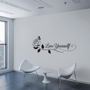 Op de afbeelding: Zwart-witte wanddecoratie met een roos en de tekst "Love Yourself". De decoratie is op een witte muur in een kamer met twee witte stoelen en een witte tafel.