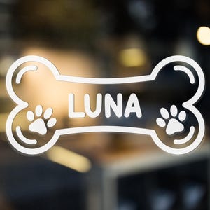 May include: A white bone-shaped decal with the name "LUNA" in white capital letters. Two paw prints are on either side of the name. The decal is likely for a pet and is displayed on a glass surface.