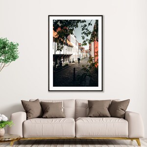 May include: A black and white photograph of a narrow street in a European city. The street is lined with old buildings, some of which are painted in bright colors. The street is cobblestone and there is a single person walking down the street.