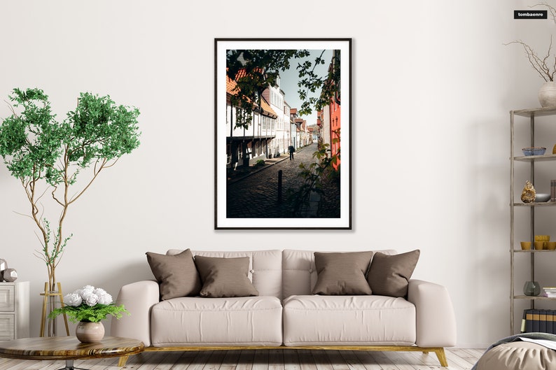 May include: A black and white photograph of a narrow street in a European city. The street is lined with old buildings, some of which are painted in bright colors. The street is cobblestone and there is a single person walking down the street.