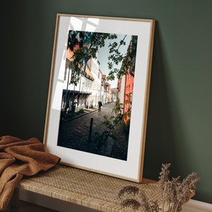 May include: A framed photograph of a narrow street in a European city. The street is lined with old buildings, and there is a single person walking down the middle of the street. The photo is in a light wood frame.