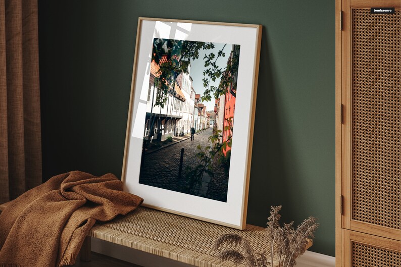 May include: A framed photograph of a narrow street in a European city. The street is lined with old buildings, and there is a single person walking down the middle of the street. The photo is in a light wood frame.