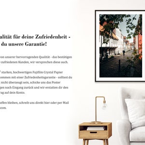 May include: A framed photograph of a narrow, cobblestone street lined with historic buildings. The buildings are a mix of white and red brick, with some featuring decorative timber framing. The street is empty except for a single person walking in the distance. The photograph is displayed on a white wall, with a wooden side table and a white sofa in the foreground.